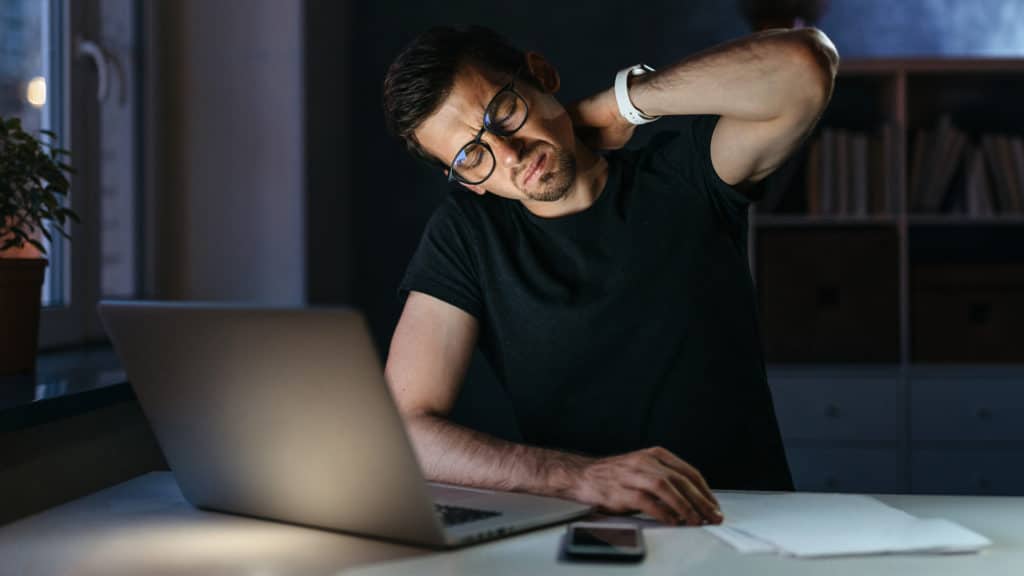 homem branco com camiseta preta, sentado em em uma mesa com um notebook cinza, levando a mão à sua nuca com expressão de dor e desconforto muscular